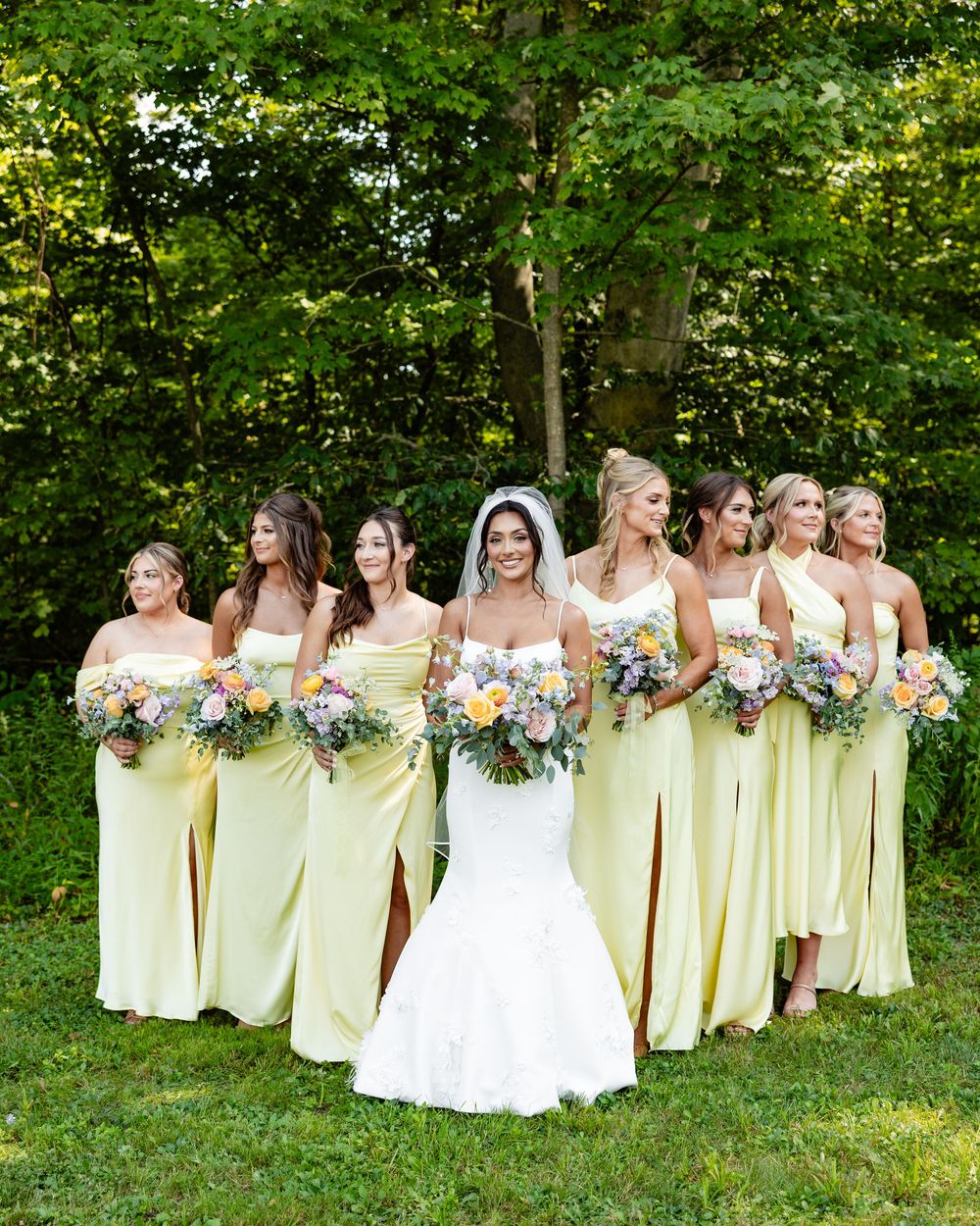 Bride in white gown poses with bridesmaids in yellow dresses holding floral bouquets on green lawn.