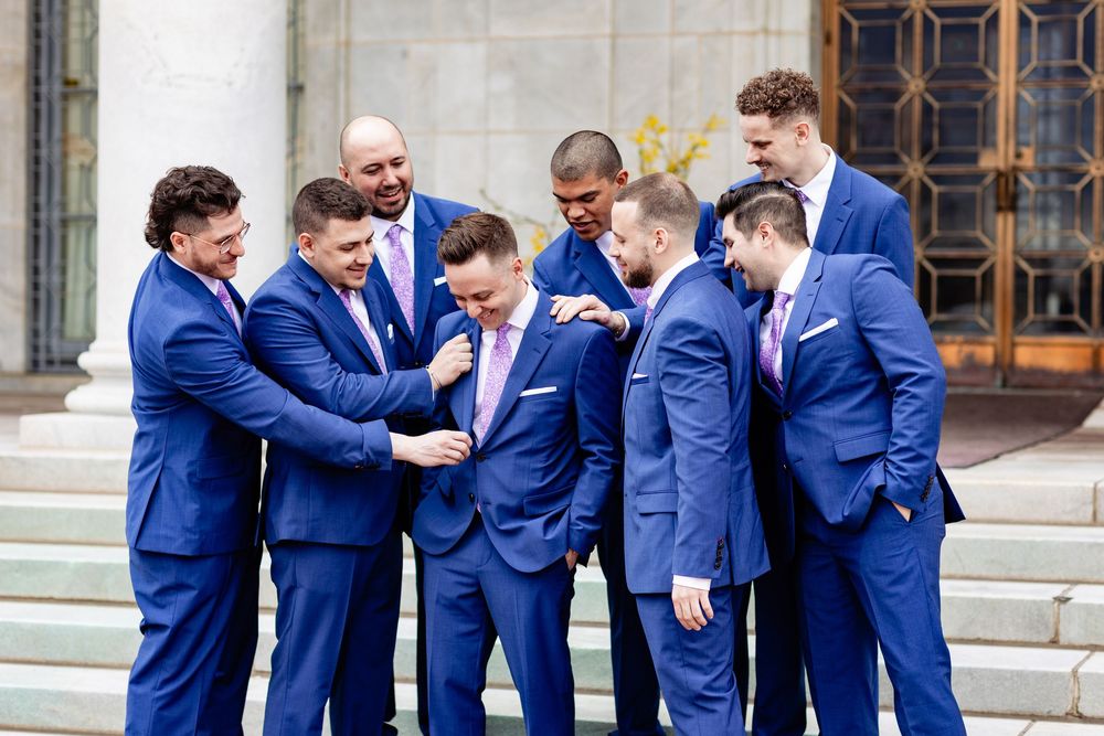 Groomsmen in matching blue suits and pink ties laughing together on stone steps during a wedding celebration.