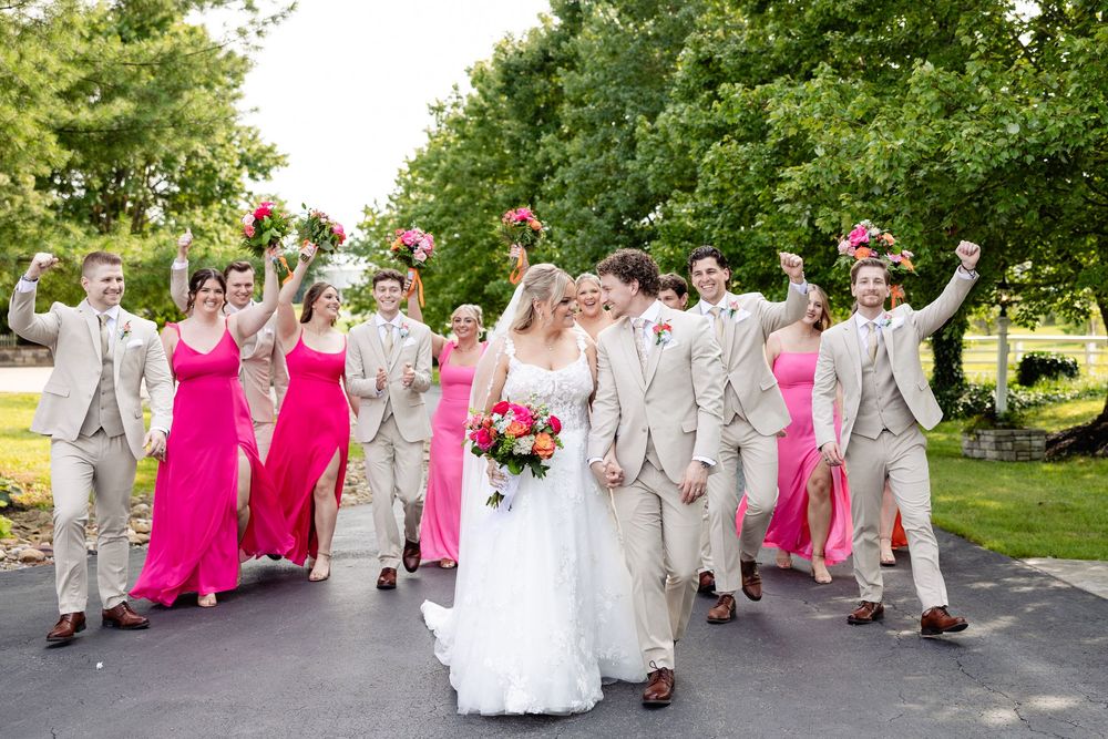 Large wedding party celebrating outdoors, bride and groom centered, bridesmaids in pink gowns and groomsmen in tan suits.