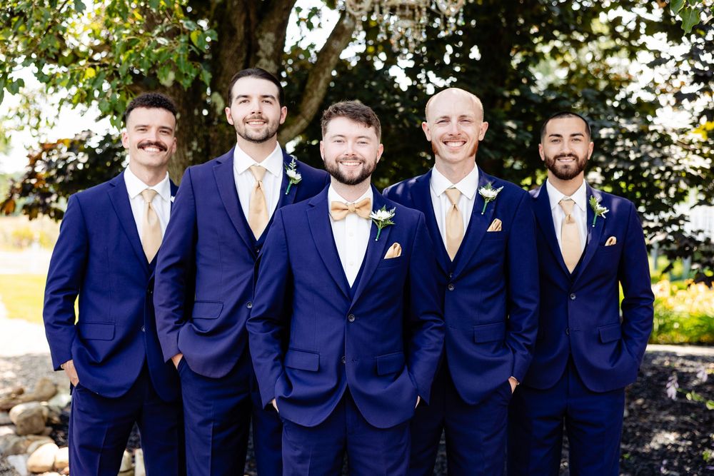 Five groomsmen in navy blue suits with gold ties pose outdoors at a wedding ceremony.