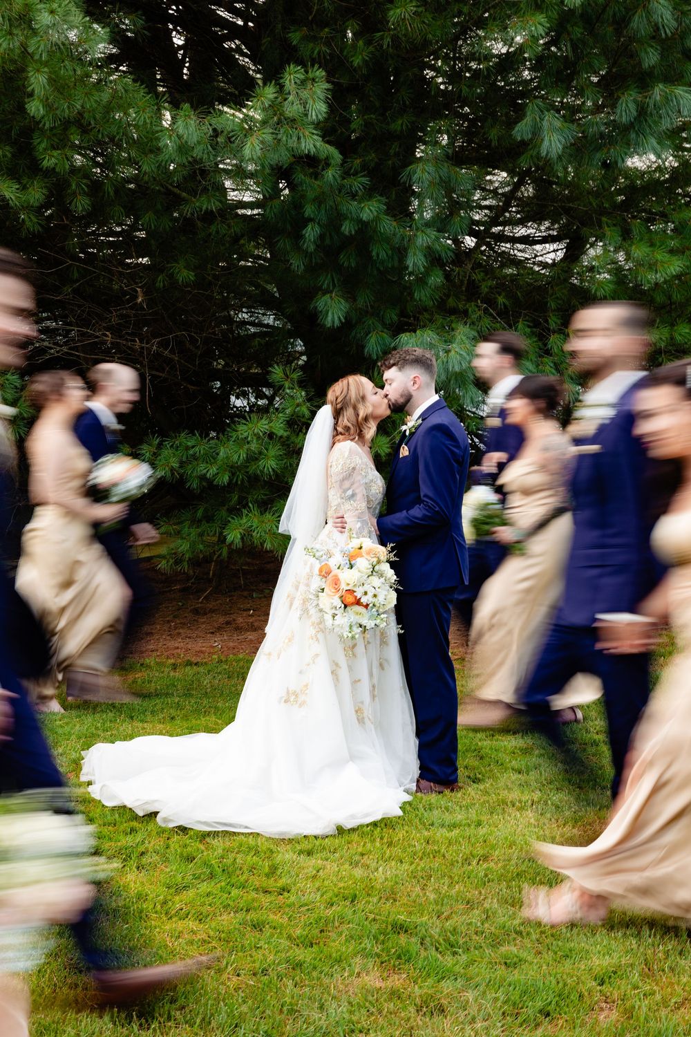 Bride and groom share a romantic moment while wedding party walks past in motion blur outdoors.