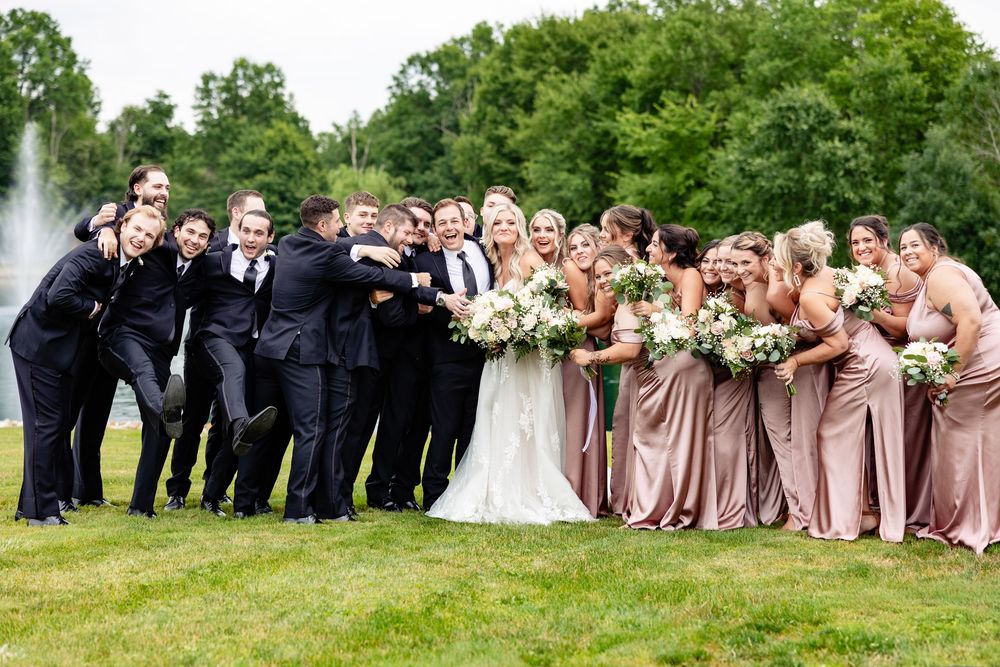 Wedding party posing outdoors, groomsmen in black suits and bridesmaids in dusty rose gowns holding white bouquets near a fountain.