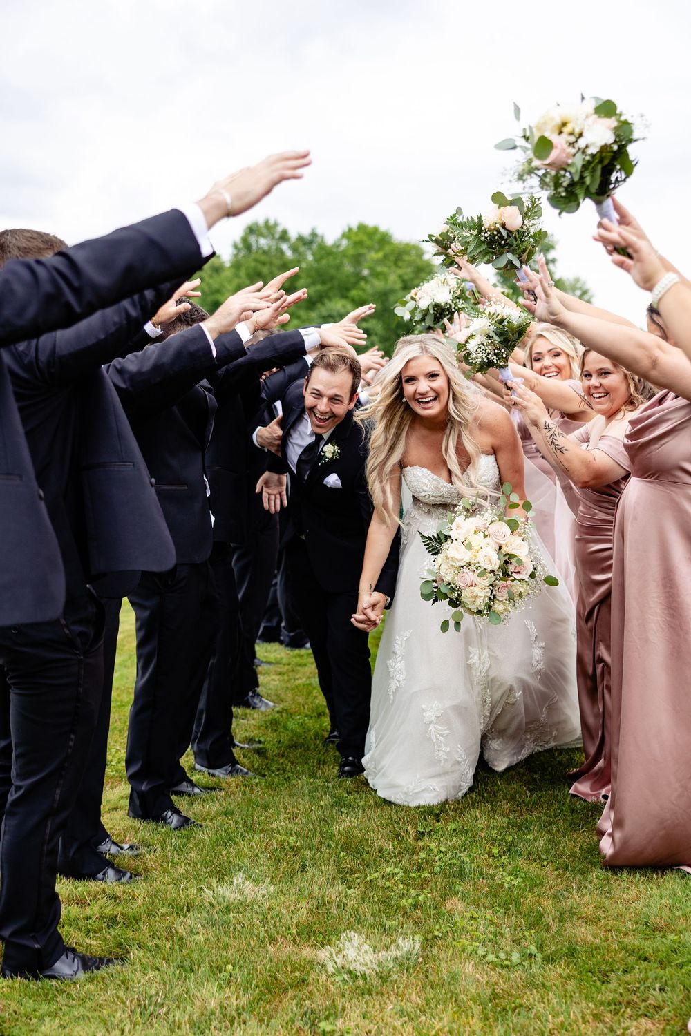 Bride and groom walking through a tunnel of raised arms formed by their wedding party on a green lawn outdoors.