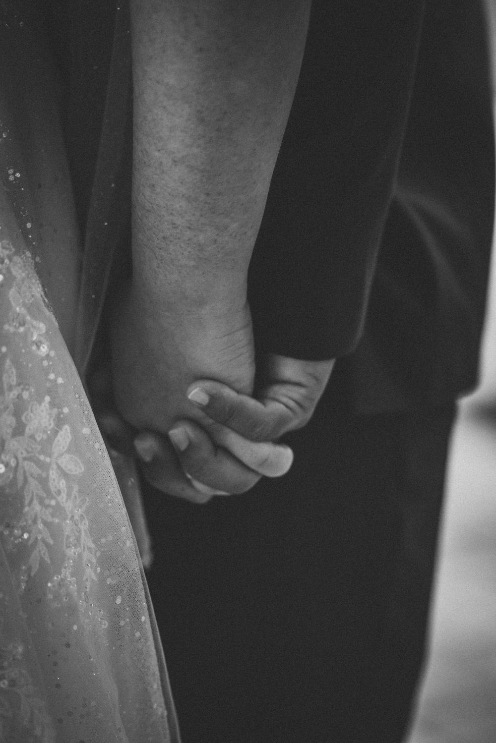 Black and white close-up of two hands clasped together, with wedding dress fabric visible.