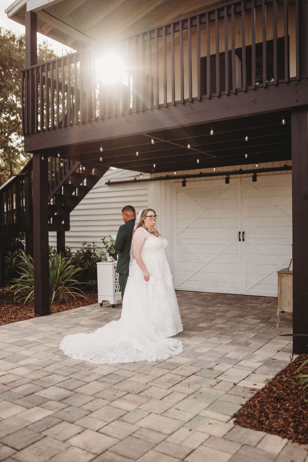 Bride and groom pose for a first look photo outside a white barn with string lights and stone patio.