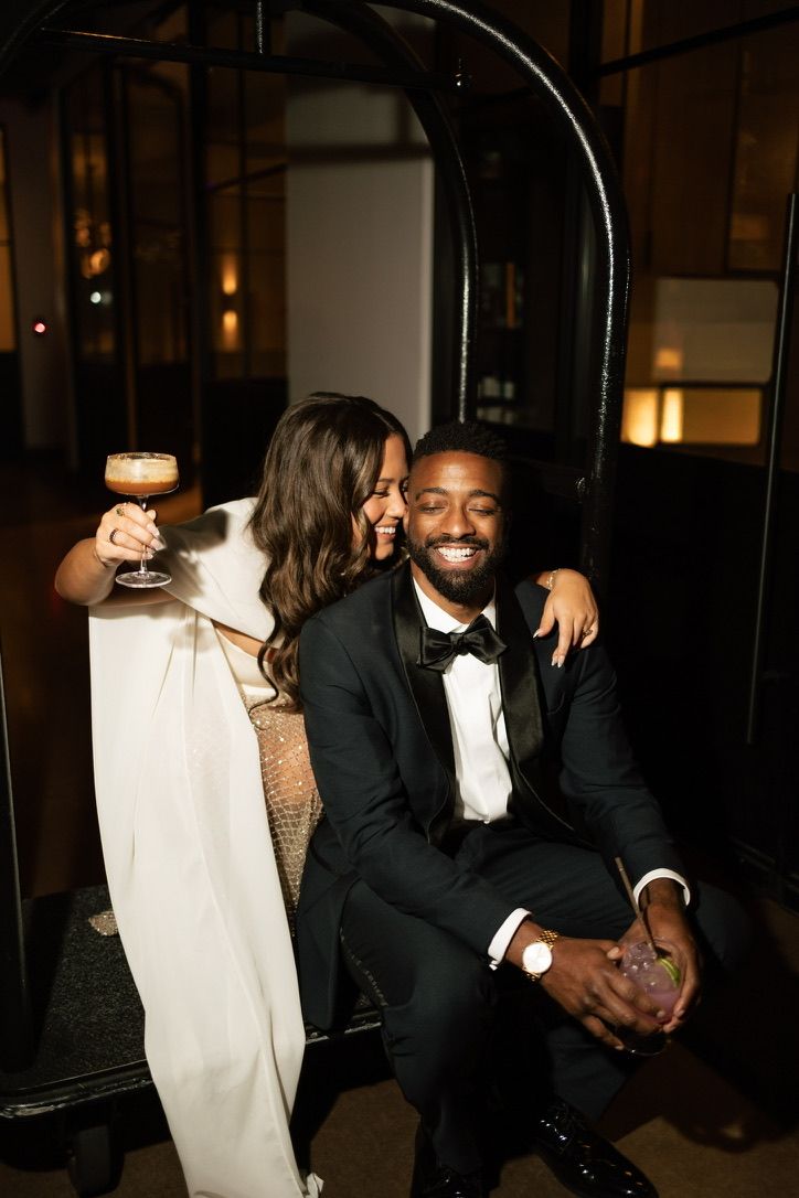 Happy couple at wedding reception, bride in white cape gown holding espresso martini, groom in black tuxedo smiling.