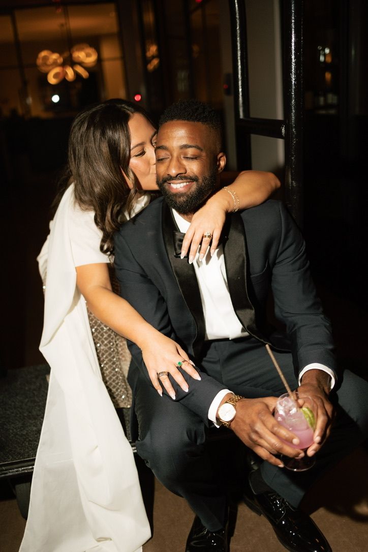Bride kissing groom on the cheek as he smiles holding a cocktail, both in wedding attire at reception.