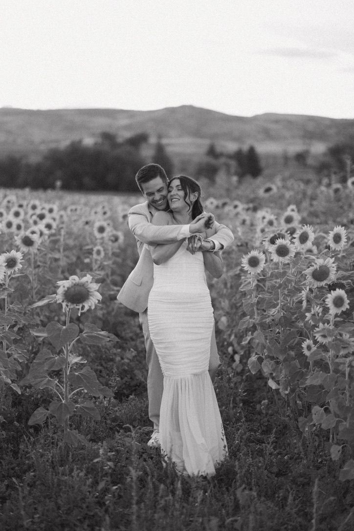 Black and white photo of a couple hugging joyfully in a sunflower field in wedding attire.