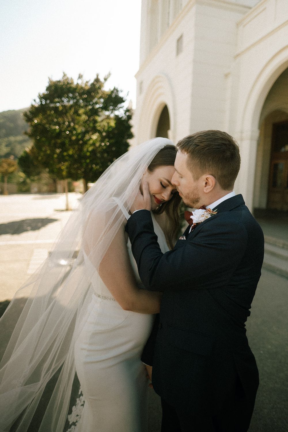 Groom in navy suit tenderly kisses bride wearing cathedral veil outside a white stone church.