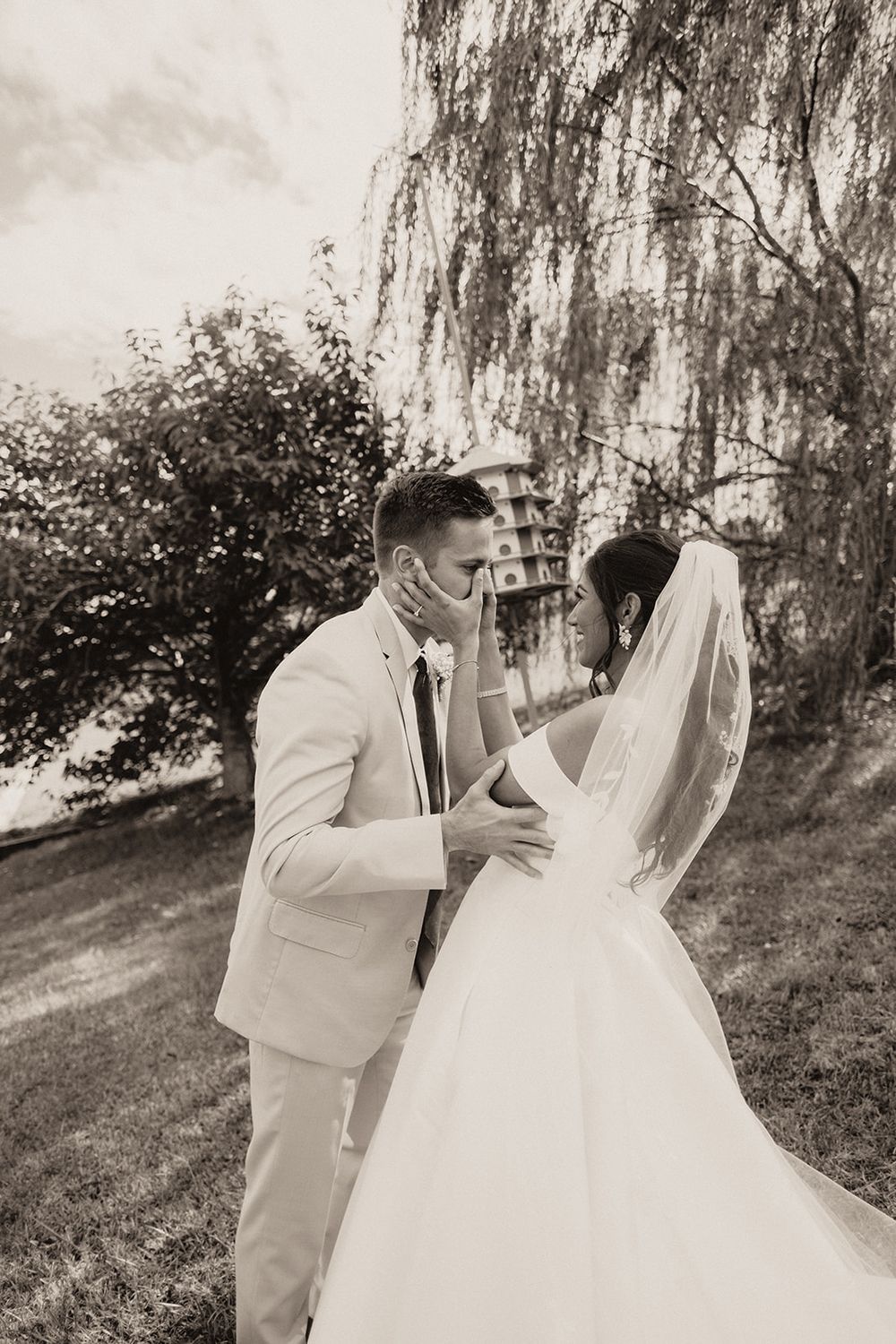 Groom in cream suit gently holds brides face during outdoor first look near weeping willow tree.