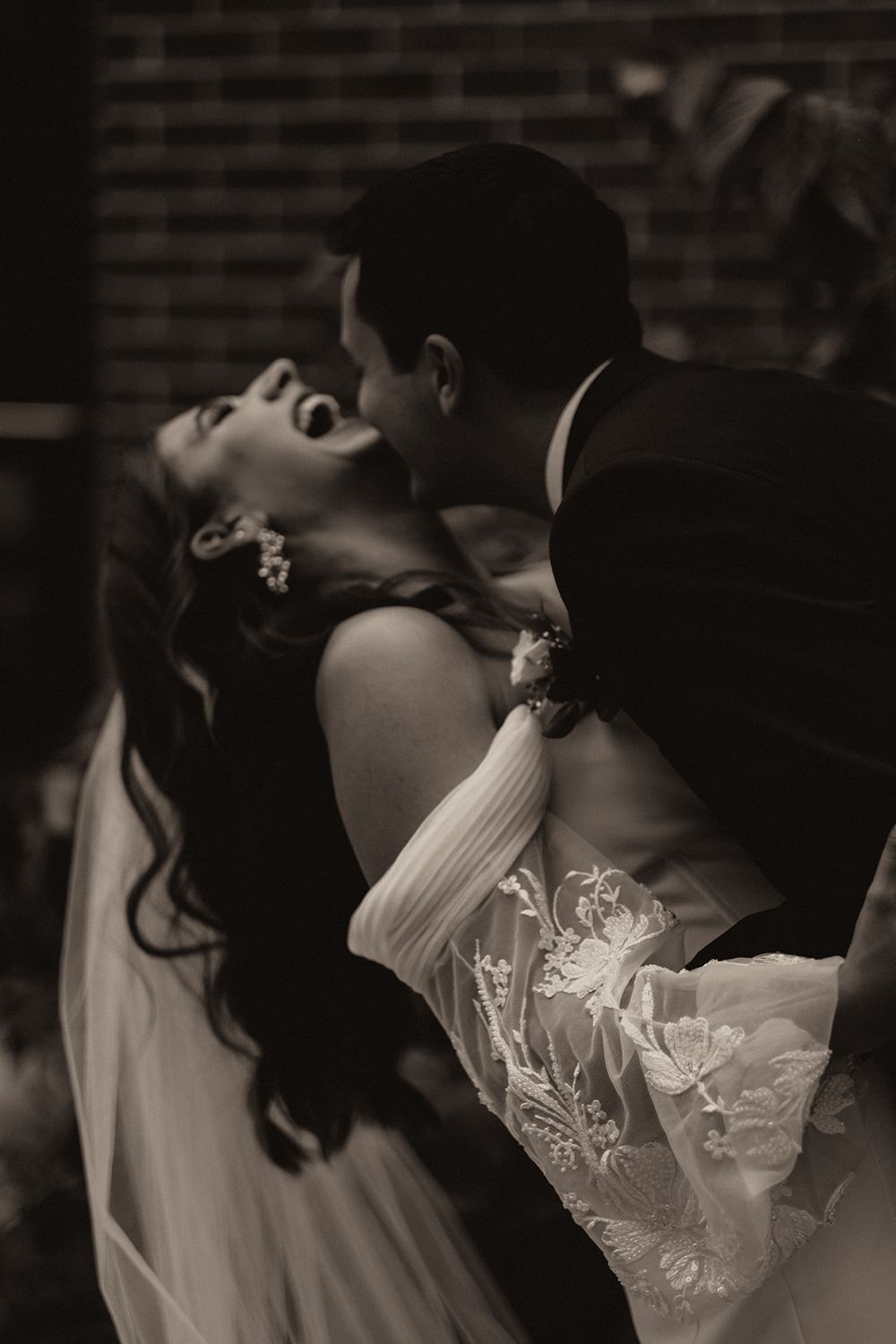 Black and white romantic photo of groom dipping laughing bride in lace wedding gown during outdoor celebration.