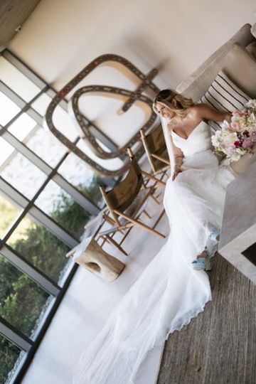 Bride in flowing white gown holds pink bouquet while leaning against wall in rustic venue interior.