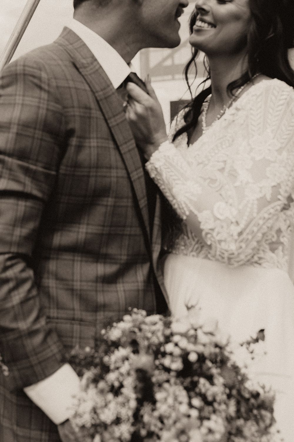 Black and white photo of smiling bride in lace gown holding bouquet while groom in plaid suit leans in.