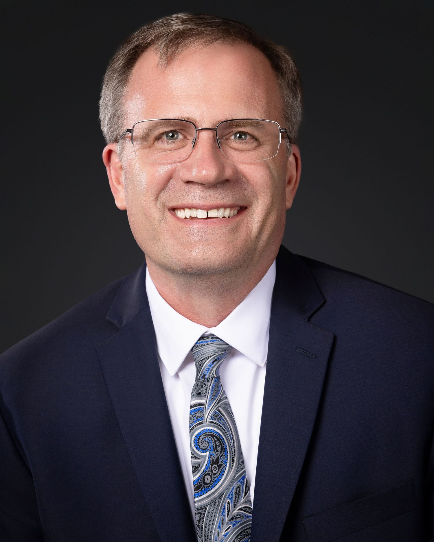 Professional headshot of businessman in navy suit with paisley tie against dark background.