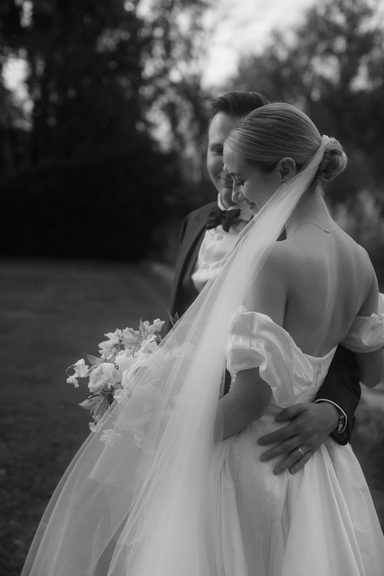 Joyful bride and groom celebrate outdoors on green lawn, groom in black tuxedo, bride in white ball gown wedding dress.