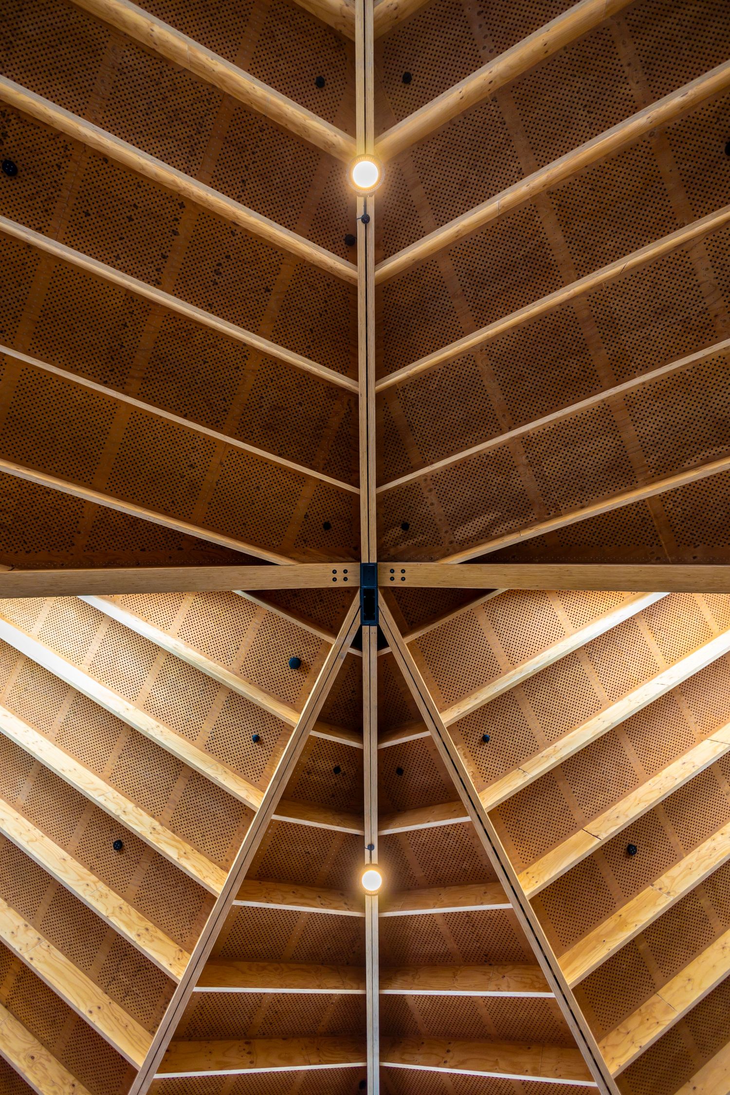 Symmetrical view looking up at a vaulted wooden ceiling with warm lighting and exposed beams forming a triangular pattern.