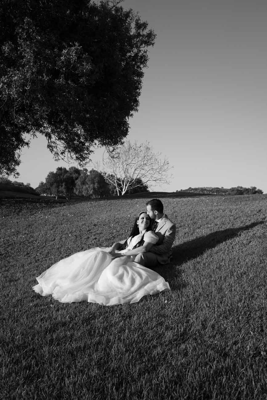 Black and white photo of a bride and groom sitting together on a grassy field, sharing a romantic moment.