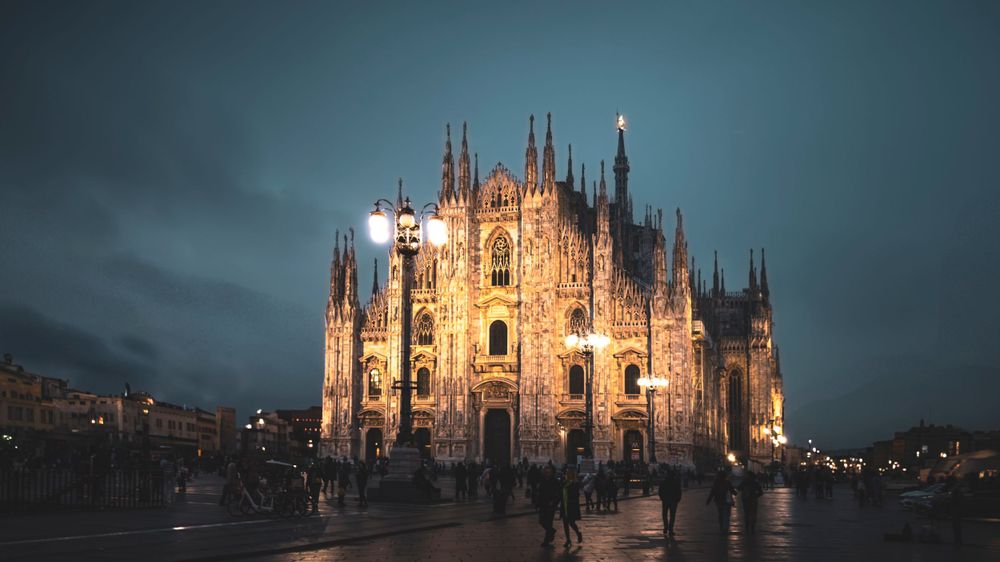 Milan Cathedral illuminated at dusk with dramatic sky and crowds gathering in the plaza.