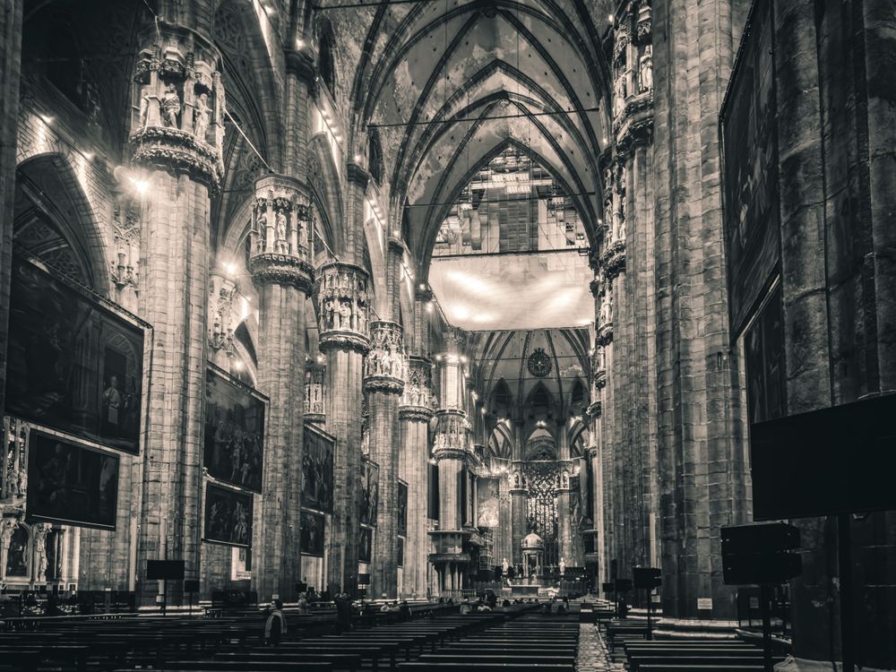Historic cathedral nave featuring gothic arches, stone columns and wooden pews in monochromatic tones.