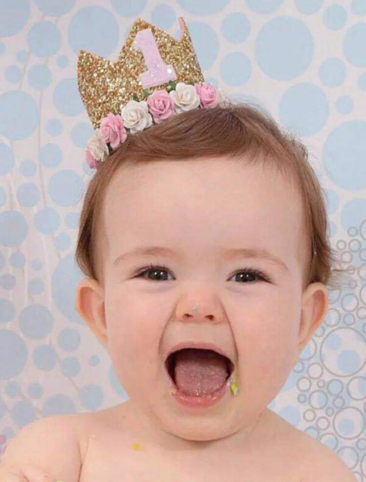 Baby wearing gold crown and floral accessories against blue patterned background.