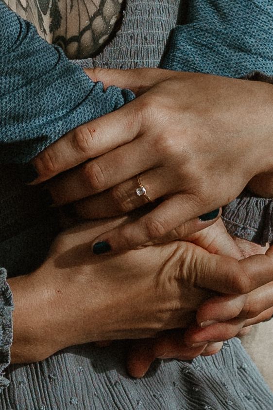 Close-up shot of hands clasped together on grey fabric with a small engagement ring sparkling in natural light.