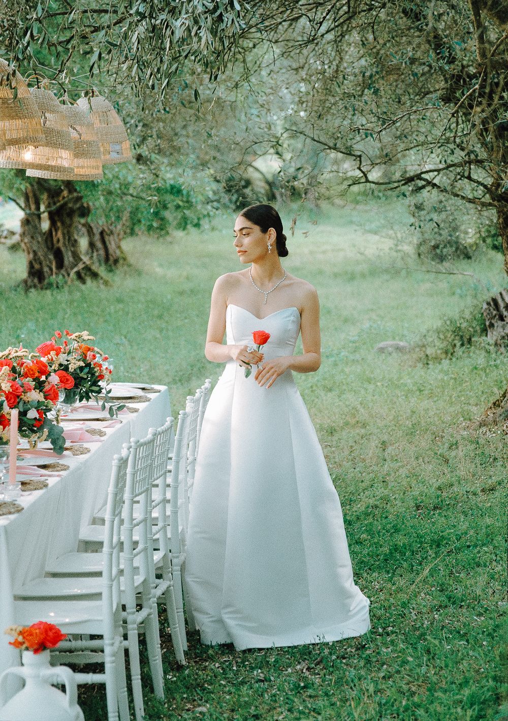 Bride in white dress standing beside an outdoor wedding table with red flowers and candlelight
