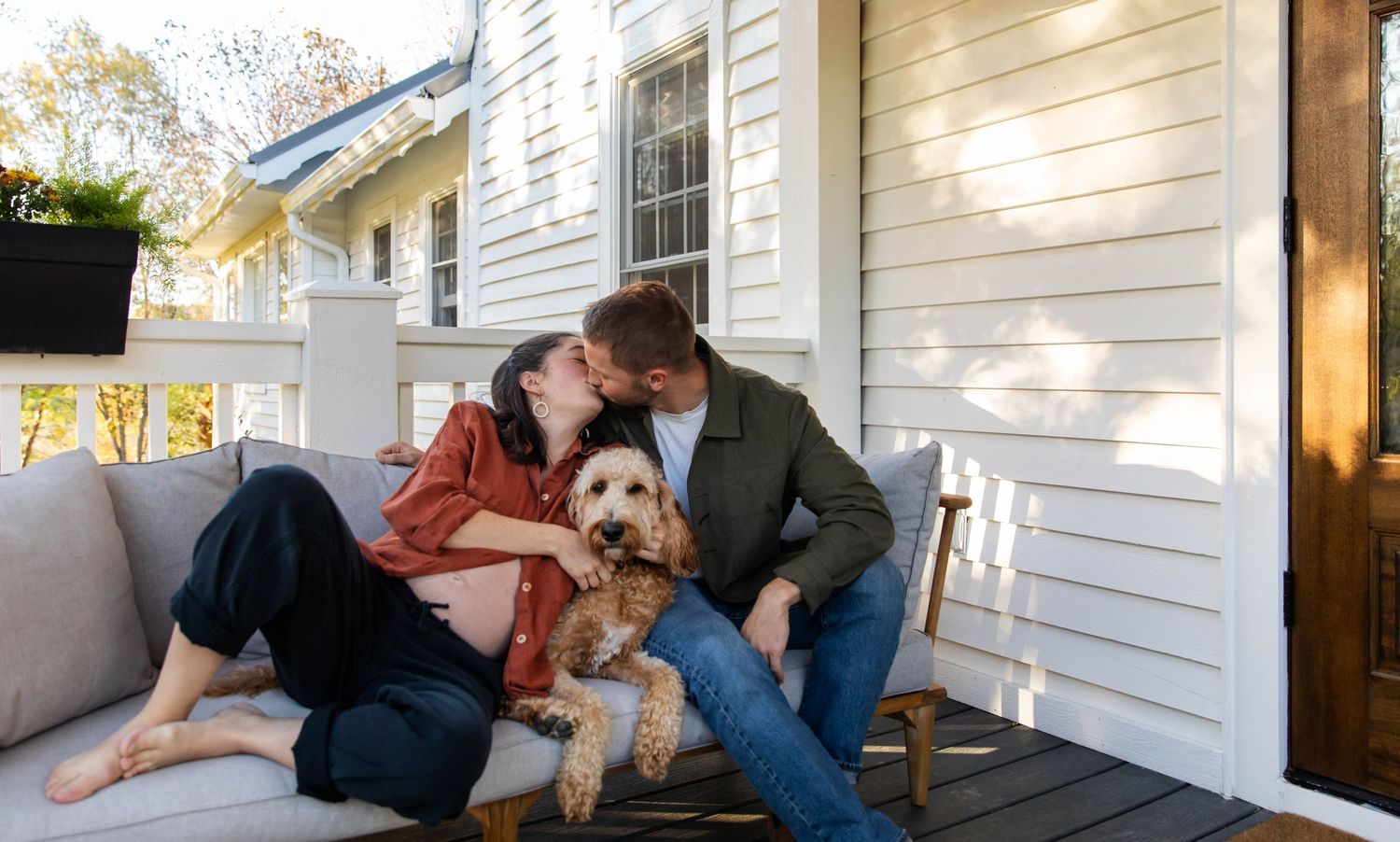 Photo of a couple sitting on their porch with their dog. She is pregnant, and they are kissing each other, during a maternity photo session