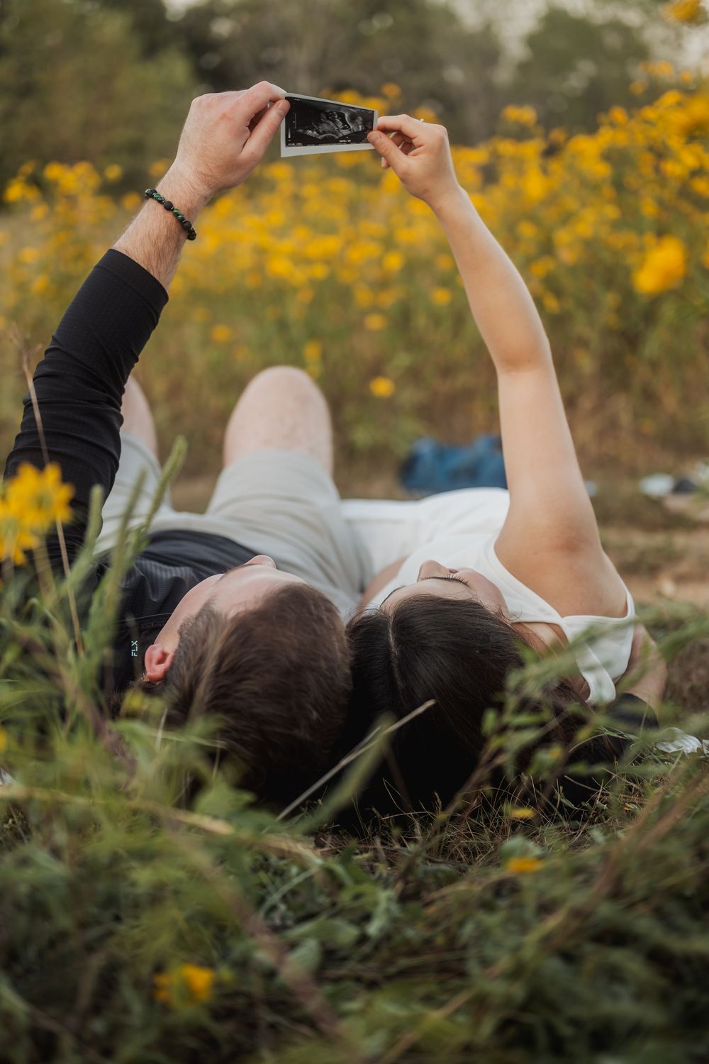 Couple viewing ultrasound photos while lying in field of yellow flowers.