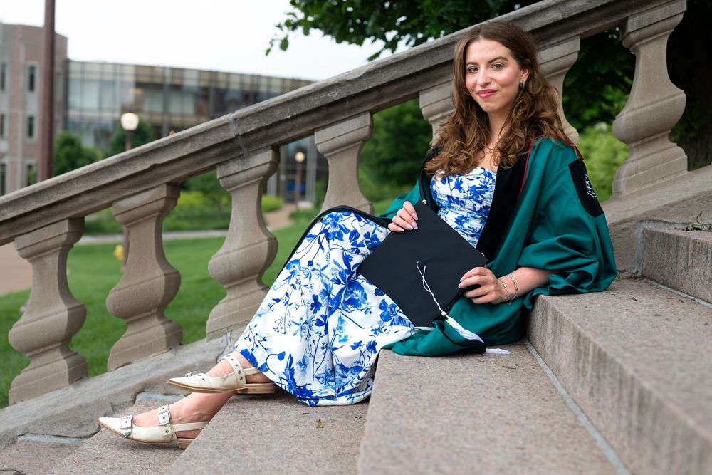 Student in floral dress and green graduation gown reclines elegantly on stone steps.