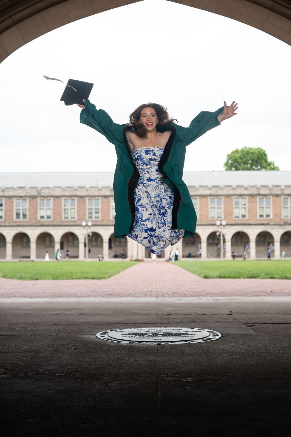 Graduate leaps joyfully under historic archway with graduation cap in hand.