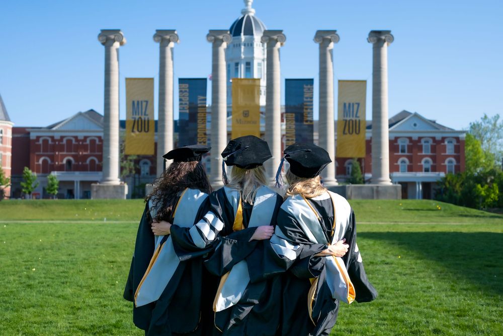 Group of graduates gather on sunny lawn in front of columned university building.