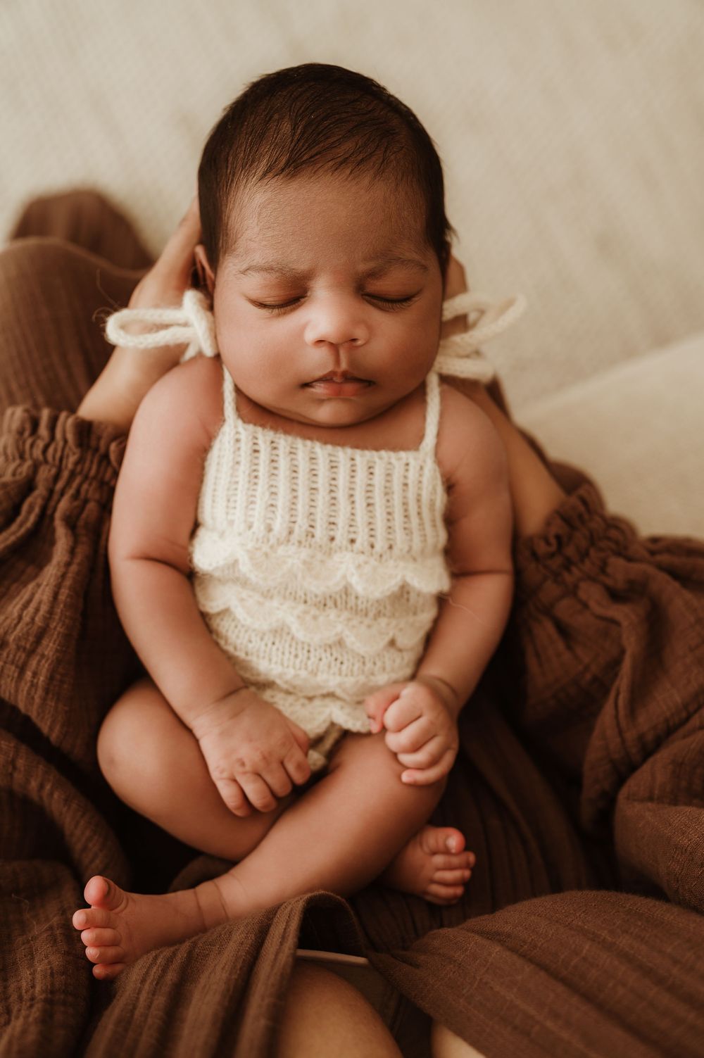 Sleeping newborn baby wearing white knit romper rests peacefully on brown blanket in warm natural light.