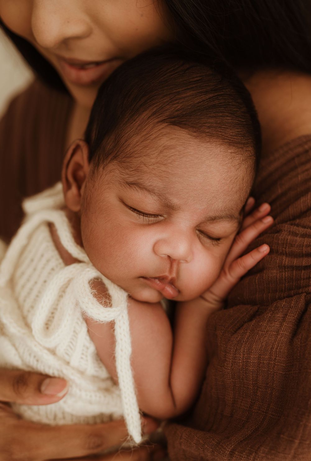 A peaceful sleeping baby wearing a white knit sweater rests against a brown fabric surface.