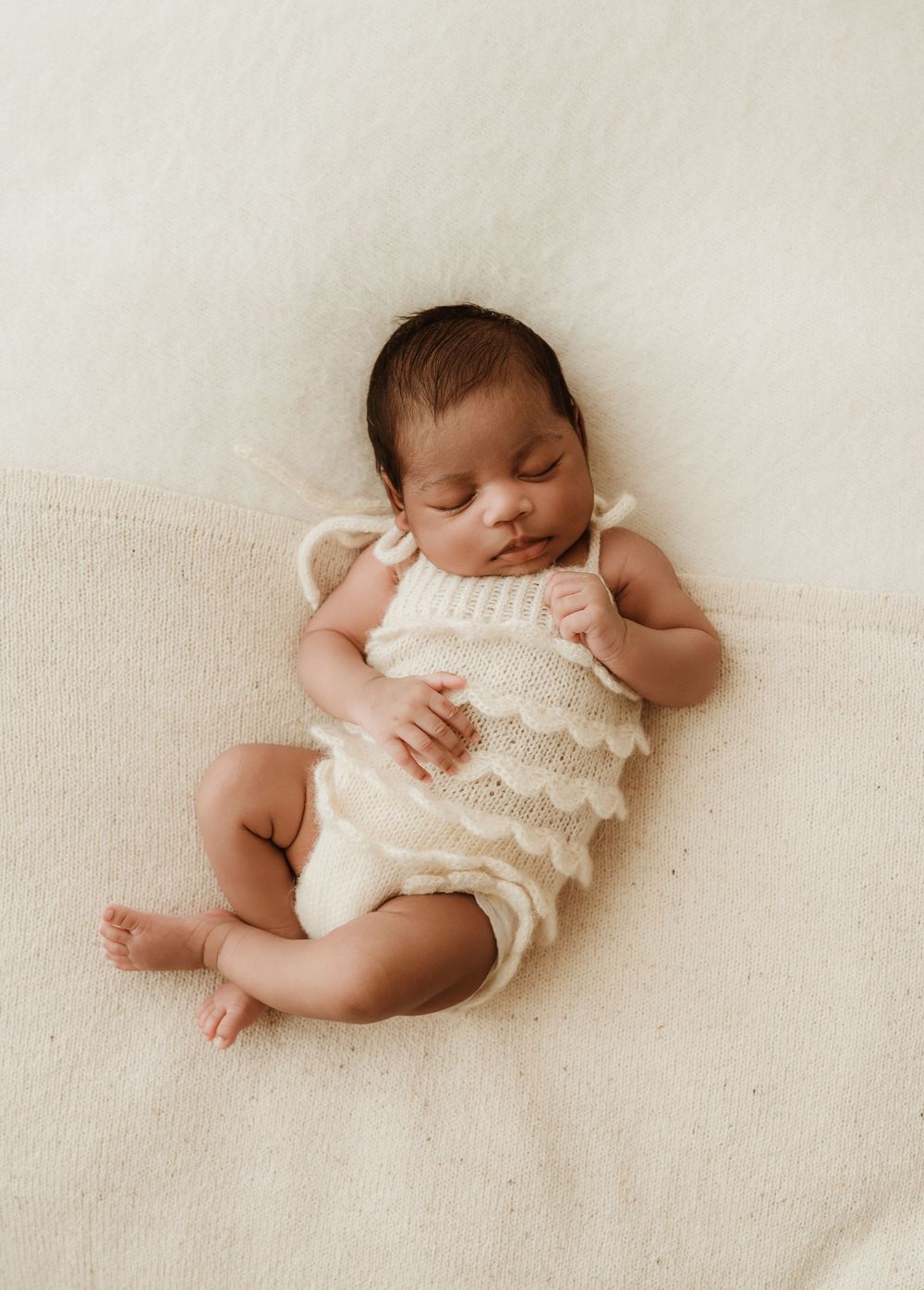 Sleeping newborn baby wearing white knit romper lying on cream colored backdrop during professional photoshoot.