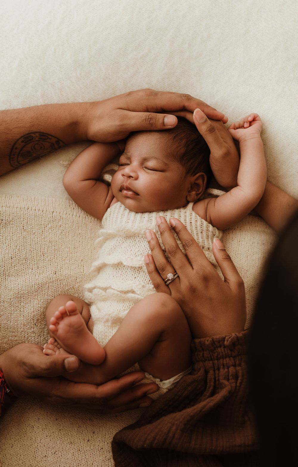 A sleeping baby wearing white lace clothing lies peacefully on a beige textured surface as gentle hands provide comfort.