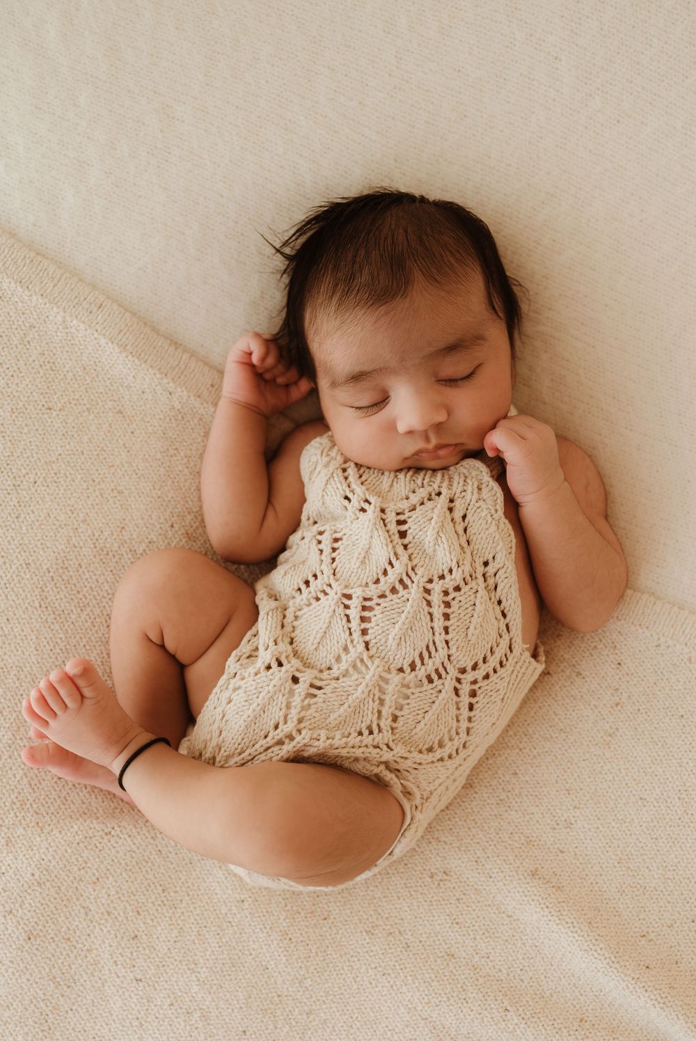 Sleeping baby in cream knit romper lying on soft beige blanket in natural light setting.