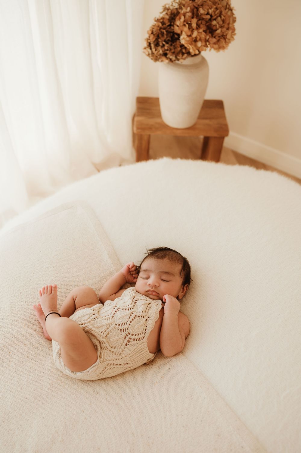 Newborn baby wearing cream knit romper sleeping on soft white blanket with floral decor in background.