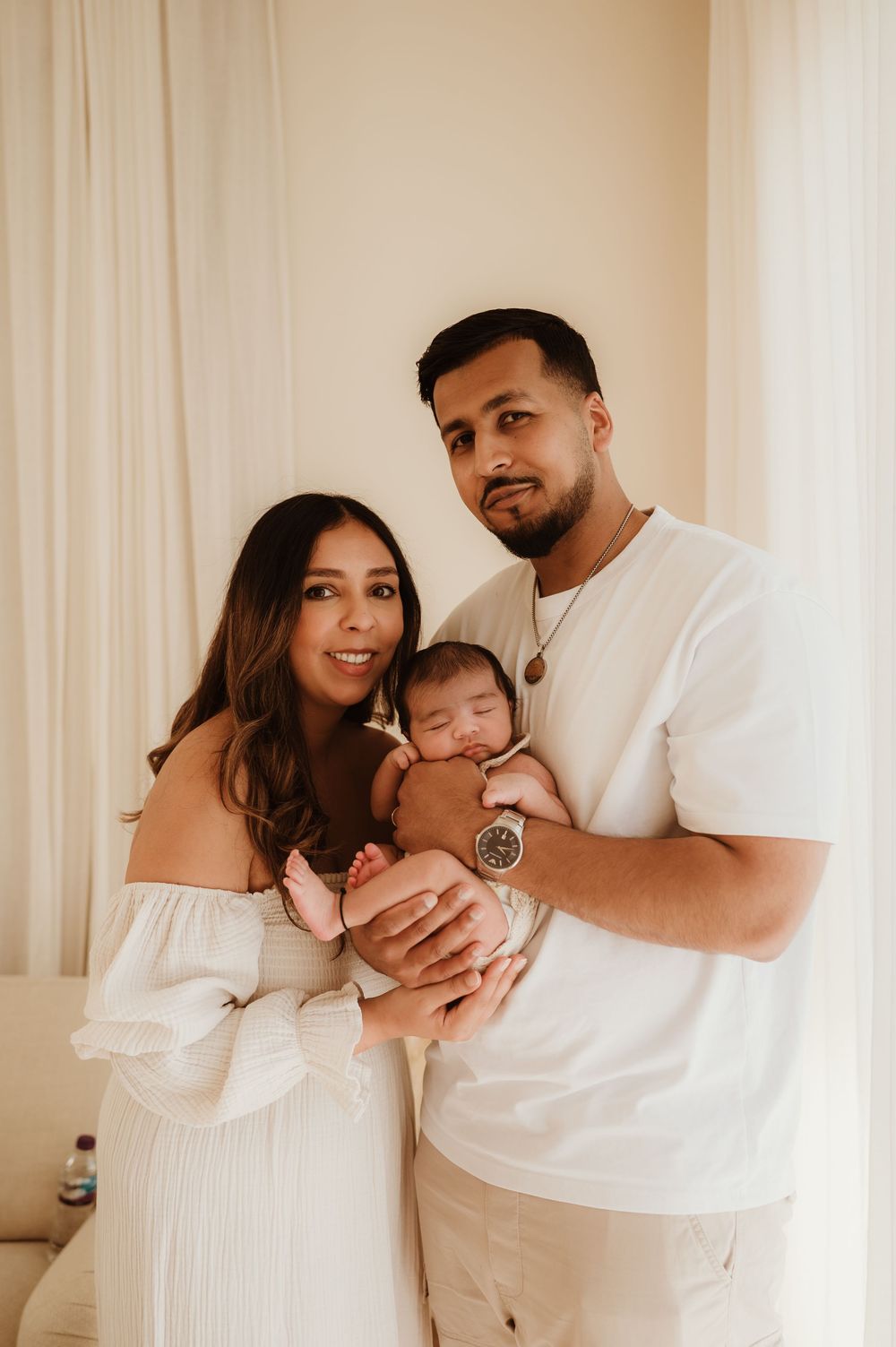 A family wearing white poses together holding their young baby in a minimalist room with curtains and warm lighting.