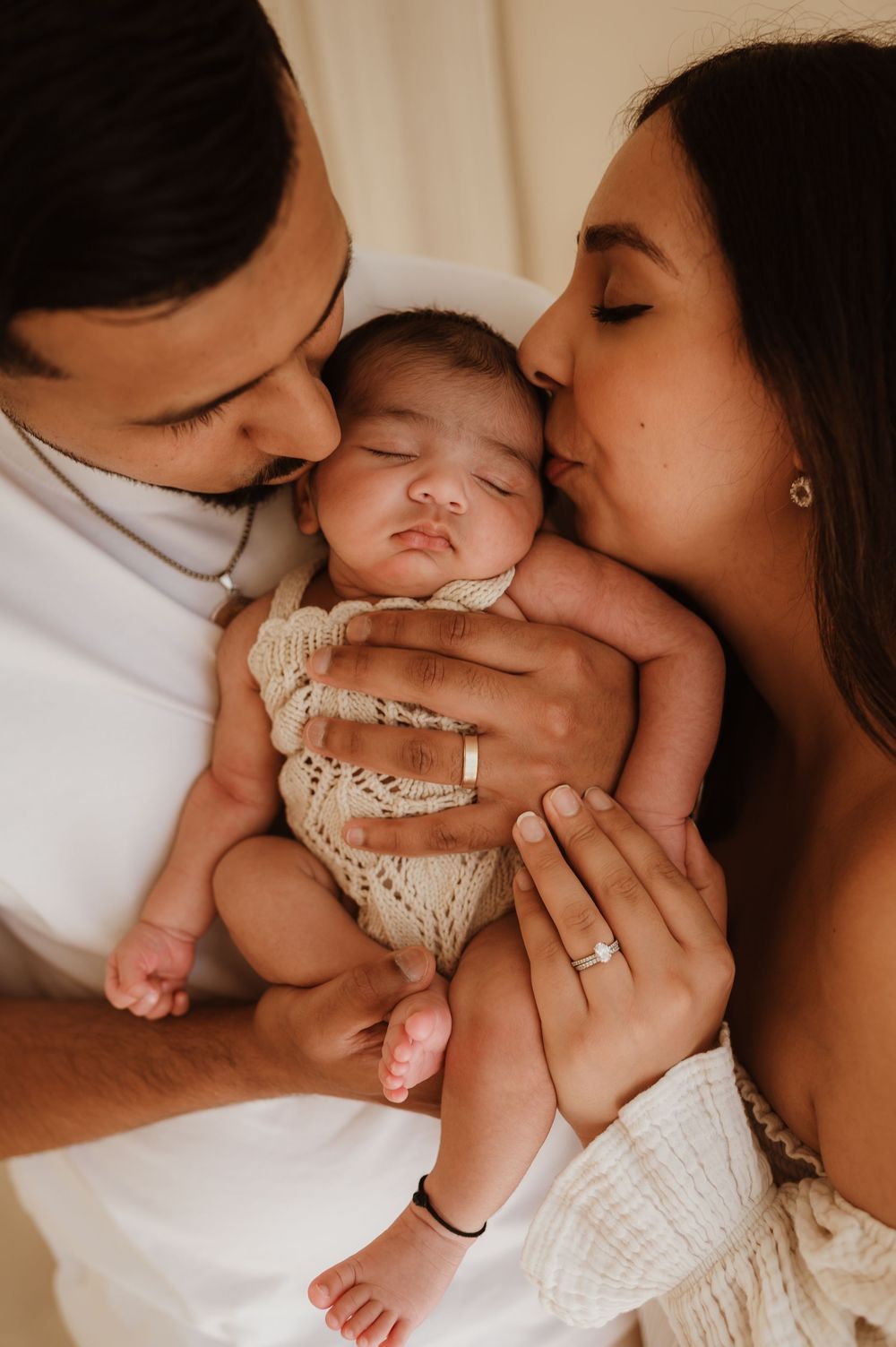A tender moment captured as parents share loving kisses with their sleeping baby in white lace clothing.