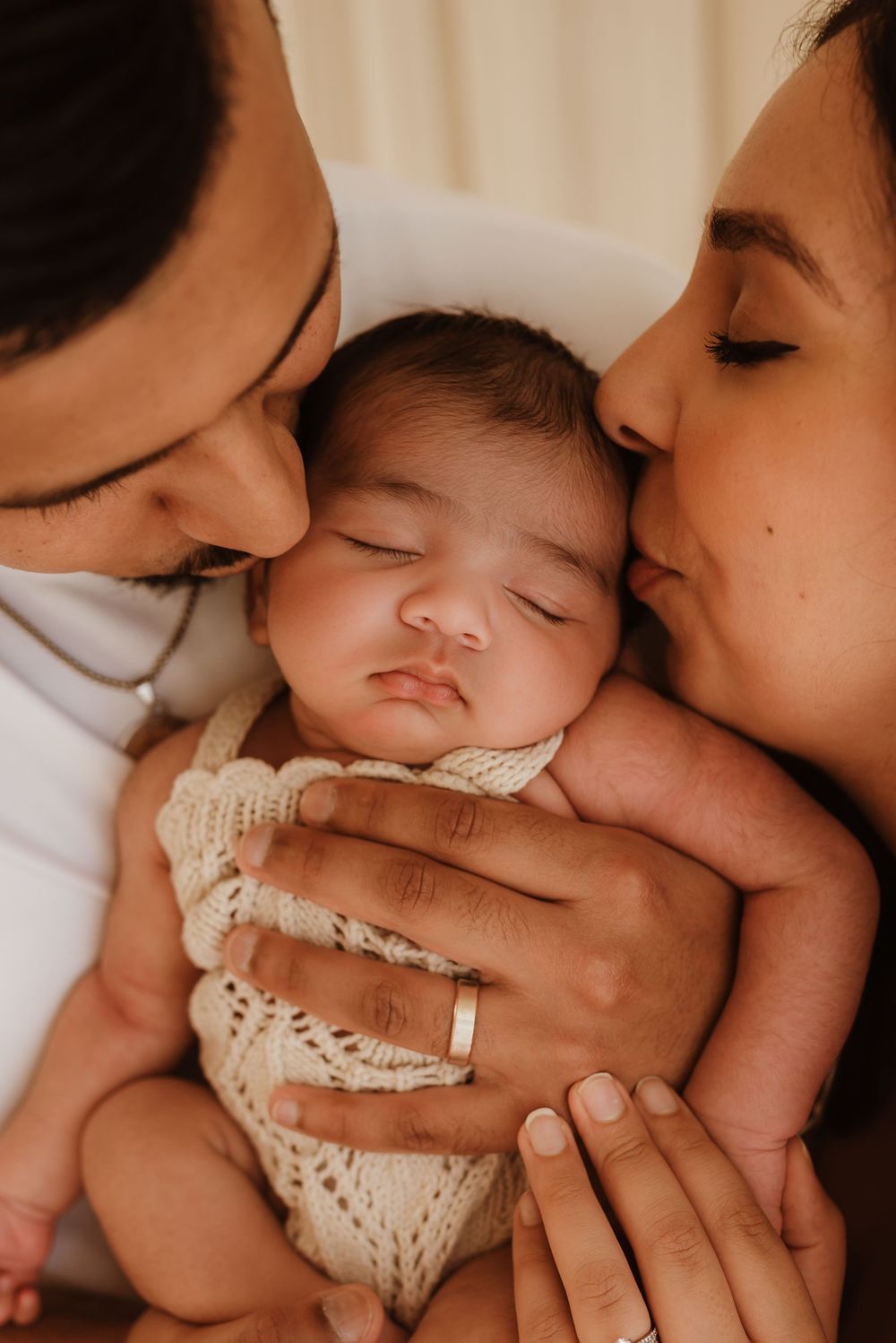 A sleeping baby in a knit romper being lovingly embraced and kissed by parents in an intimate family portrait.