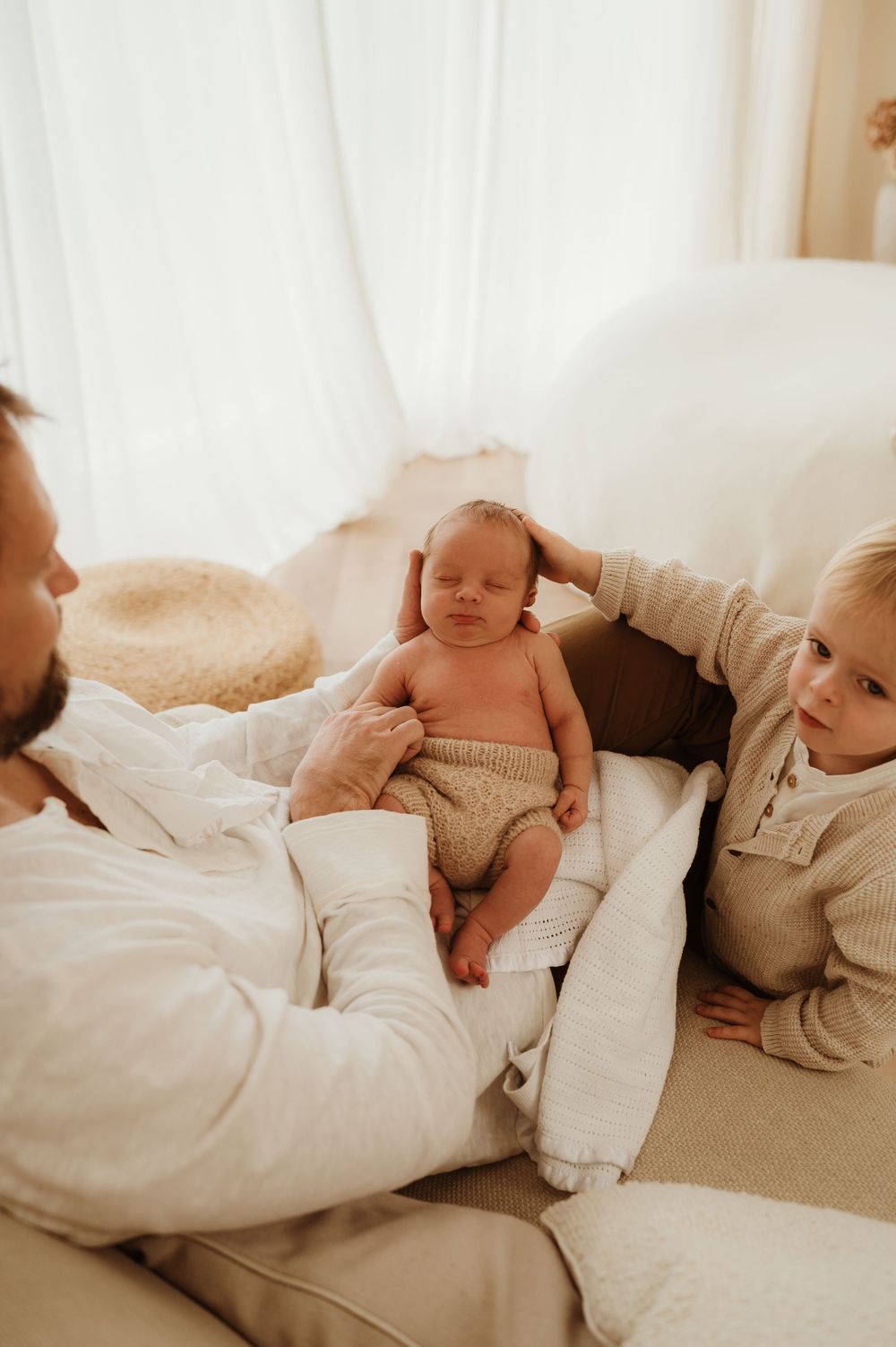 A family relaxes together on a couch, with a sleeping newborn baby in a diaper nestled between them.