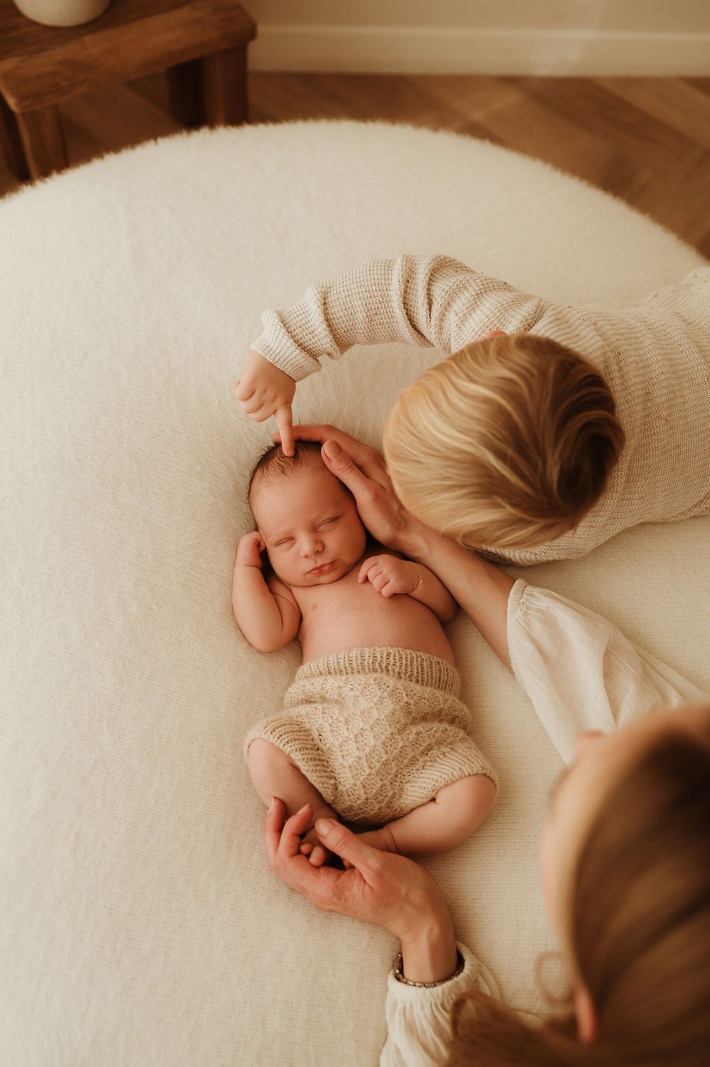 A newborn baby wearing a knit romper lies peacefully on a white blanket during a photography session.