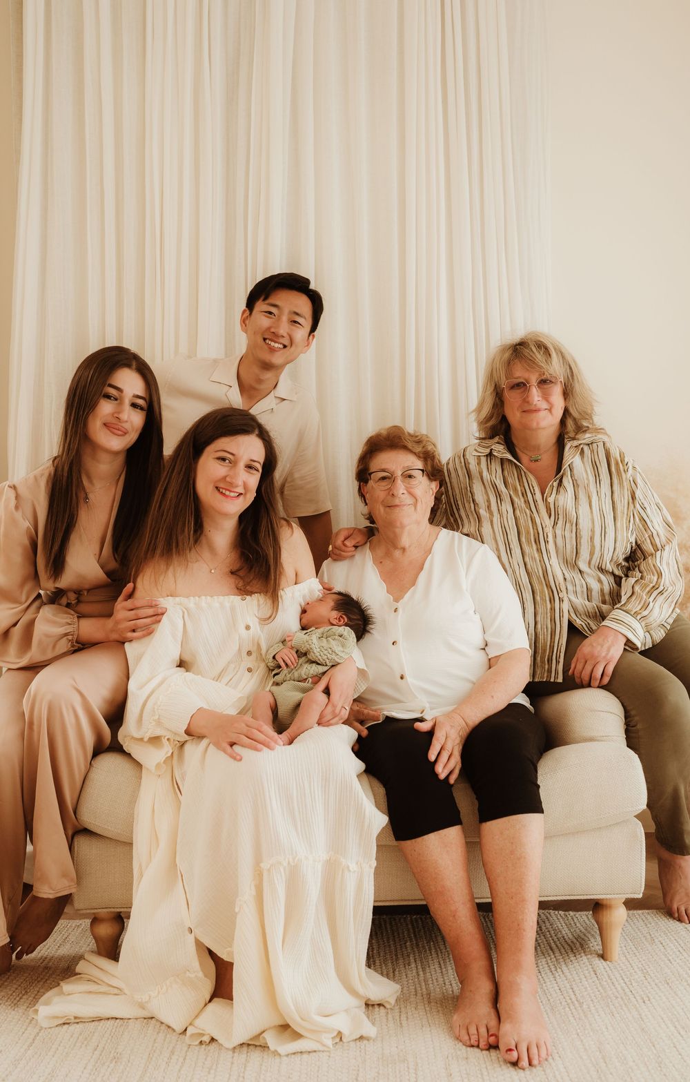 Multi-generational family with baby gathered on beige couch for intimate indoor portrait session.