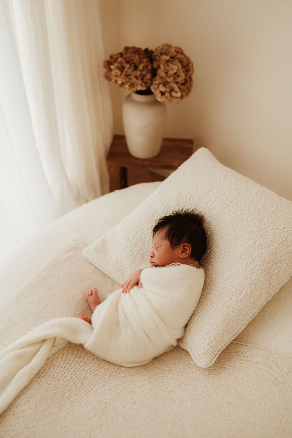 A peaceful newborn baby swaddled in white rests on a soft pillow next to a vase with dried flowers.