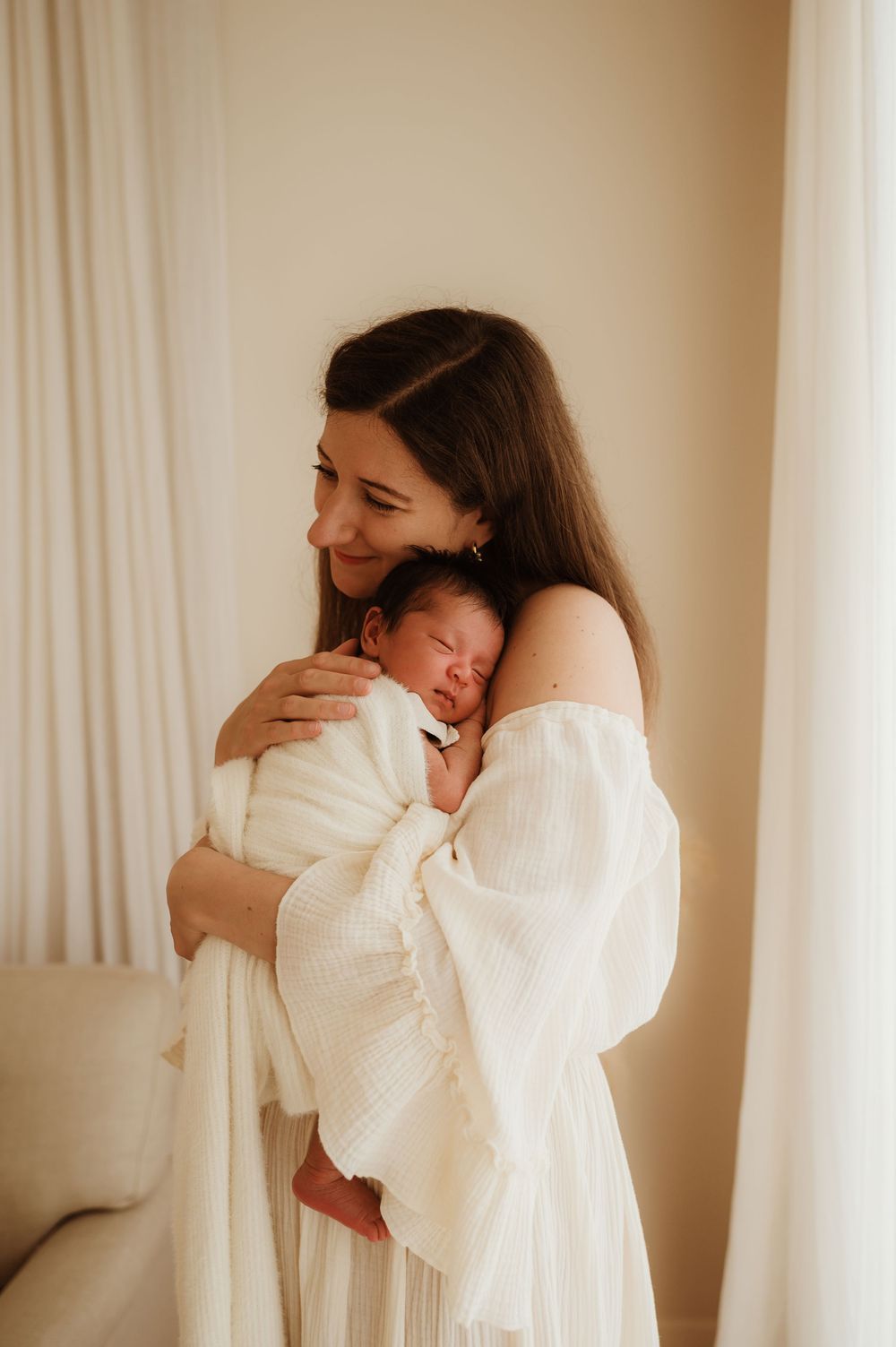 Mother in white dress holding sleeping newborn baby against soft curtained backdrop.