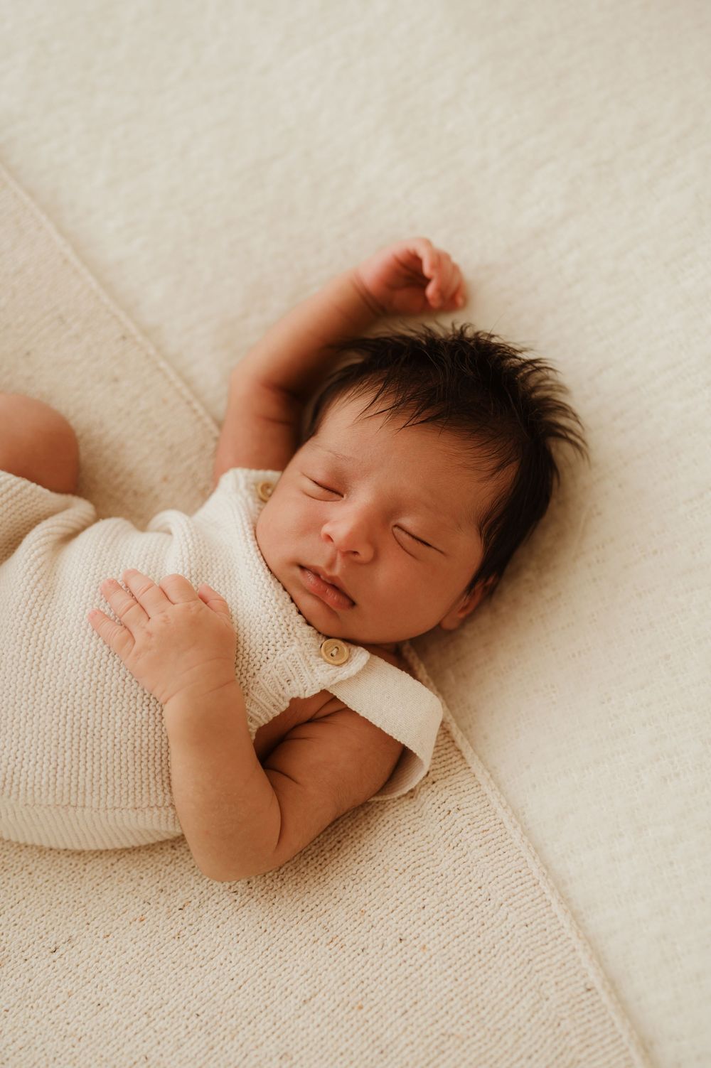 Sleeping newborn baby with dark hair wearing white knit clothing rests peacefully on cream-colored fabric.