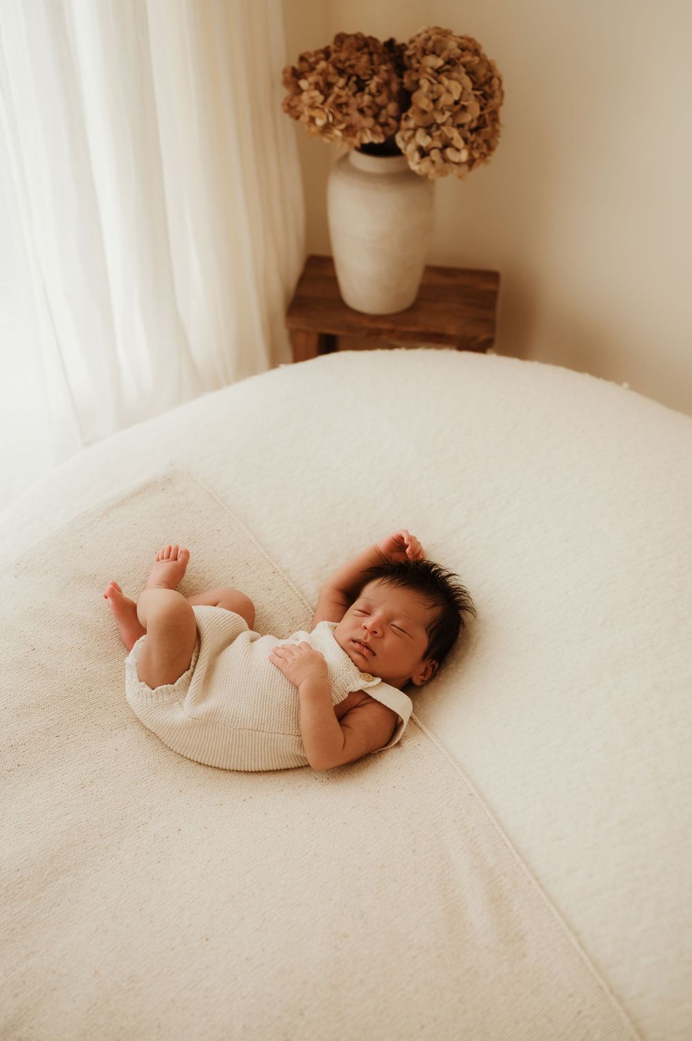 A sleeping baby lies peacefully on white bedding near a vase of dried flowers.