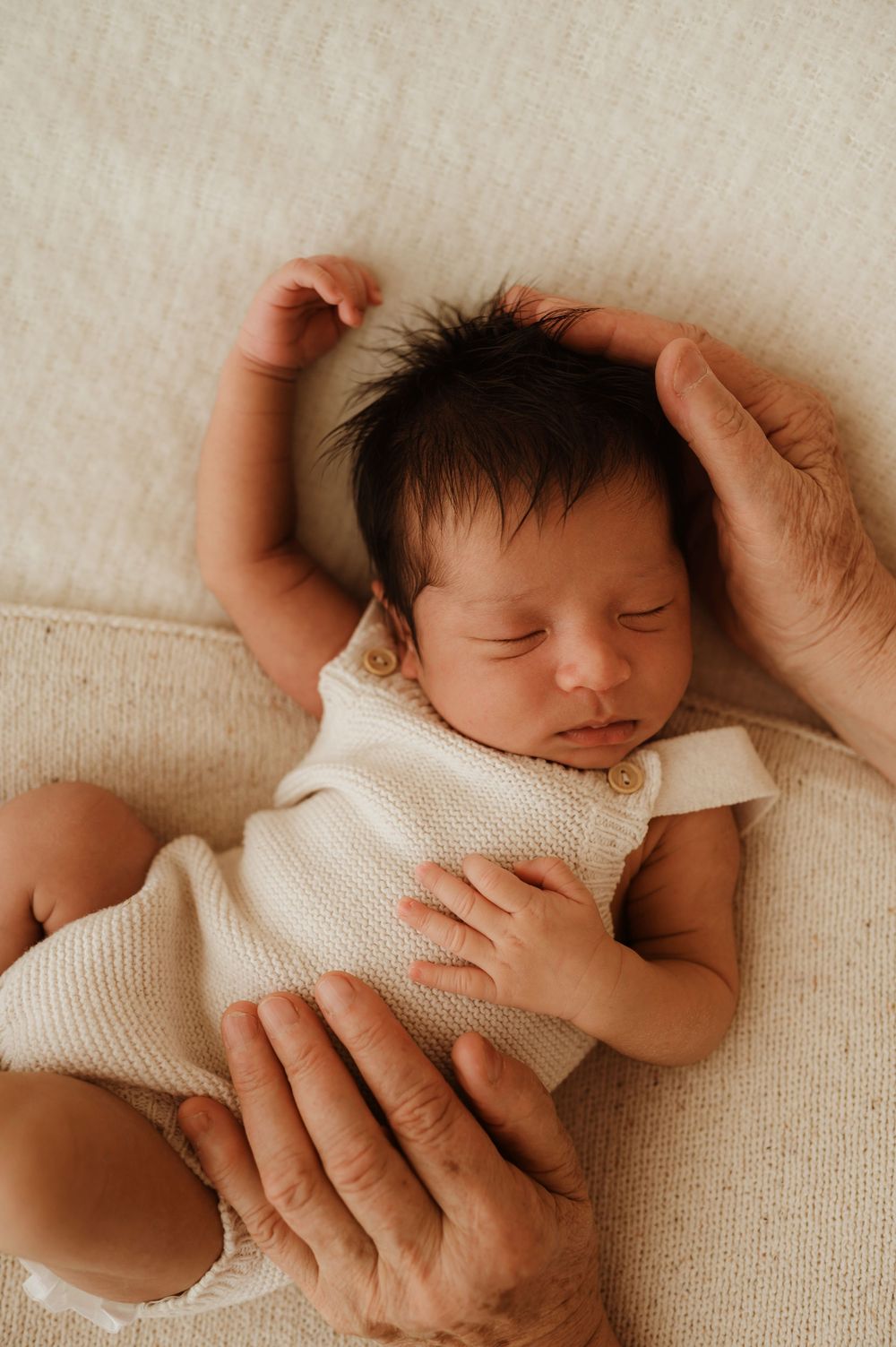 A sleeping newborn in white clothing being cradled gently by caring hands against a cream background.