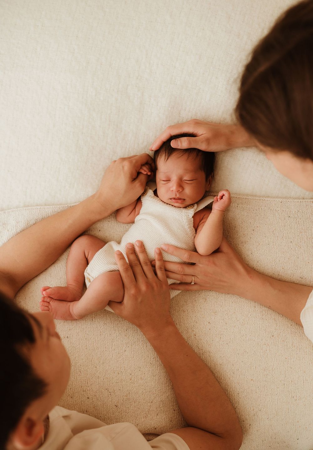 Hands tenderly cradling a sleeping newborn baby in white onesie on cream-colored couch.