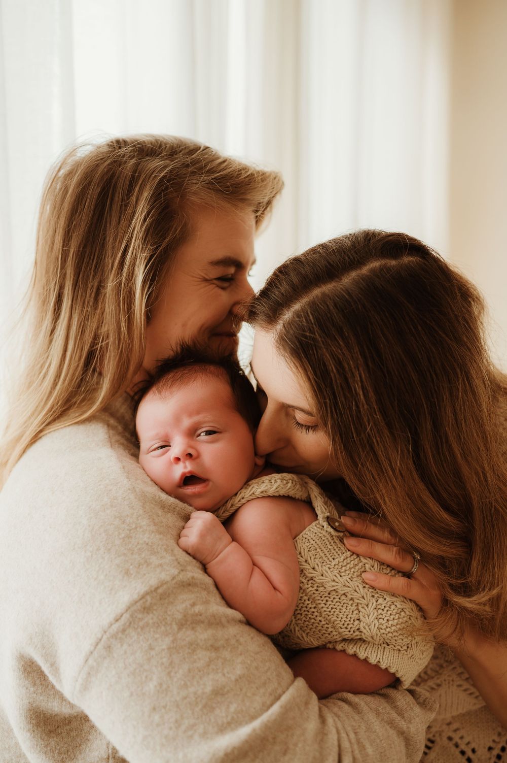 A tender family moment as parents share an intimate embrace with their newborn baby wrapped in a knit blanket.